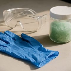 Nitrilhandskar, skyddsglasögon och en tät plastburk med gröna kristaller på en arbetsbänk